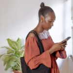 Young woman in rust-colored dress using smartphone indoors by window. Natural light and casual style.